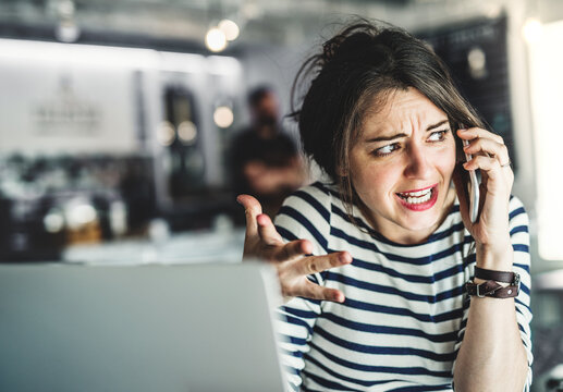 Woman working remotely from coffee shop, has annoying phone call. Cappucino and laptop on table. Female freelancer working on notebook in cafe.