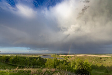  A vibrant rainbow arcs over a tranquil countryside scene, where a river meanders through lush greenery and a forested hill.