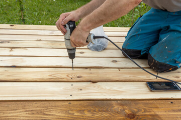 A man is using a power drill to secure a wooden plank to another piece of wood. His right hand is on the drill, his left hand is on the wood. The power cord is visible on the wood.