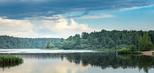 A serene lake, surrounded by a lush green forest, basks under a partly cloudy summer sky. The water is calm and reflects the sky, trees, and clouds above..