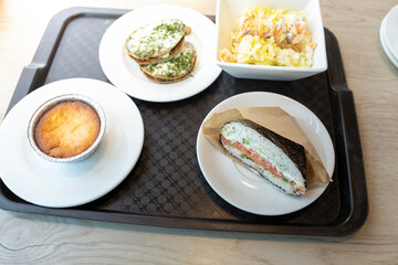 A tray of food, likely from a cafeteria or food court, features a variety of dishes. The tray contains a small baked dish, a sandwich-like dish, a stack of savory bites, and a side of salad.