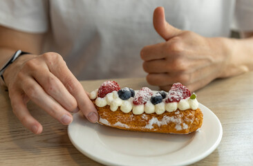 A person gives a thumbs-up while a delicious eclair with whipped cream and fresh berries sits on a...