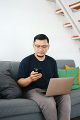 Asian man sitting on couch at home using laptop and cell phone