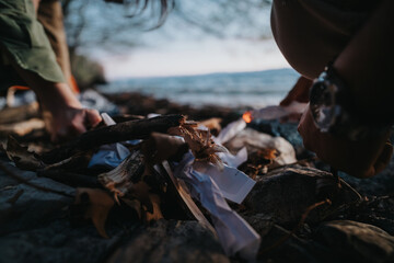 Two people gently arrange logs and paper to start a campfire by the lake at dusk, capturing the essence of outdoor adventure and camaraderie.