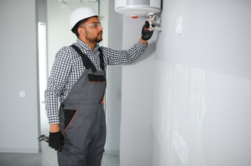 Indian plumber using screwdriver while fixing boiler or water heater, working on heating system in apartment