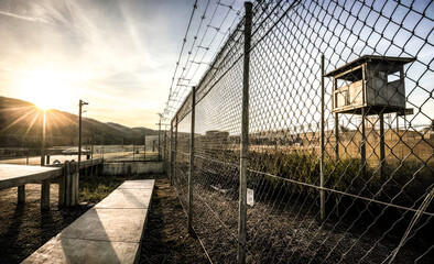 Fototapeta premium prison environment. Secure Facility Highlighted, Person Walking Concrete Path, Tall Chain-Link Fence, Barbed Wire Top, Wood Observation Tower