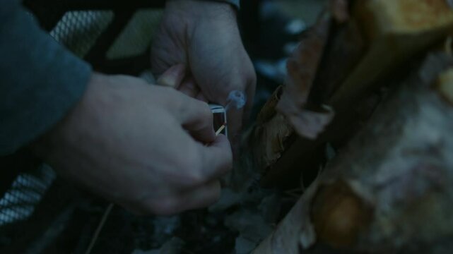 Man striking match on matchbox to light fire at base of teepee logs in pit