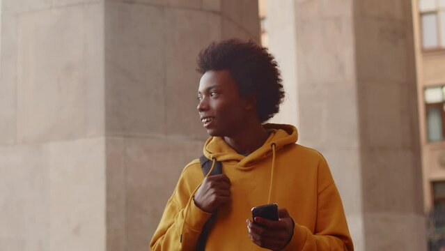 Young black programmer in casual clothes walking alongside the building outdoors, looking at the smartphone and smiling after a successful working day