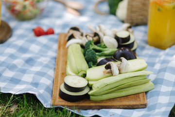 Slised vegetables on wooden board outside. Fresh vegetables outdoors on checkered napkin. Zucchini, eggplant, mushrooms and broccoli.
