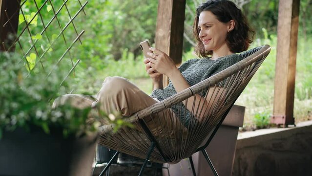 A young Caucasian woman using a smartphone while sitting in a comfy armchair on the terrace of a country house