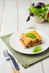 Sliced ​​Greek moussaka, layered casserole of eggplant, potatoes, minced meat with tomato sauce on a white plate on a light wooden background. Greek cuisine. Eggplant dishes.