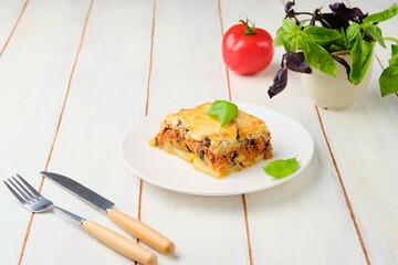 Sliced ​​Greek moussaka, layered casserole of eggplant, potatoes, minced meat with tomato sauce on a white plate on a light wooden background. Greek cuisine. Eggplant dishes.