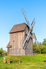 A wooden windmill stands tall against a blue sky in Borovnice, Czechia. The windmill is surrounded by lush green grass and trees.