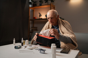 Elderly sick man sitting on couch, using tissue, wrapped in blanket, surrounded by medications on...