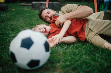 Obraz premium Dad lying on grass with son, laughing and having fun. Father and boy playing football, catching each other, falling to the ground. Fatherhood and Father's Day.