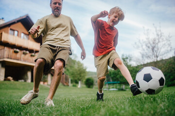 Dad having fun with young son, playing football, running and kicking the ball. Playing on a lawn in...