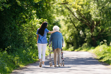 Female caregiver and senior woman with walker on walk in nature. Nurse and elderly woman enjoying a...