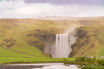 Fototapeta premium Skógafoss waterfall, Iceland, early summer, rain, cloudy, green pastures, distant people visible on the cliff sides. River in the foreground.