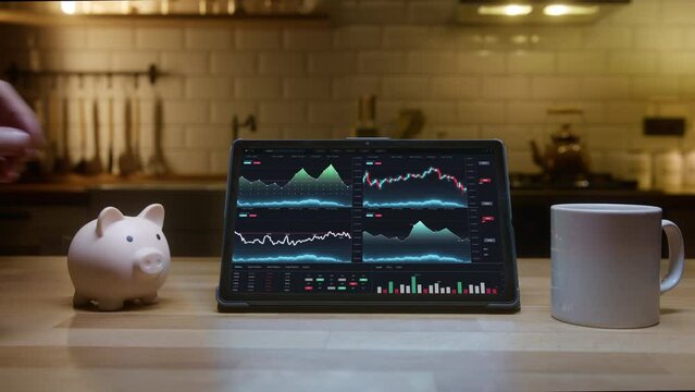 Tablet computer with financial charts on screen standing on the kitchen table at night. Unknown person puts coin money into piggy bank. Saving money and finance concept