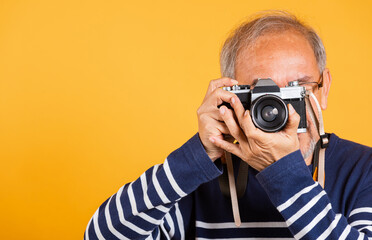Portrait Asian old man wearing glasses look viewfinder vintage SLR camera studio shot isolated yellow background, smiling happy Photographer elderly man gray haired taking a picture of himself