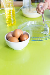 woman's hand rubbing oil with a kitchen tongue into pastry bowl. bowls with eggs and dough on pistachio green kitchen countertop.