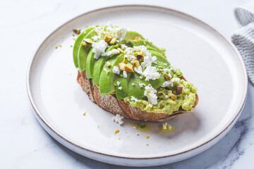 Avocado toast with feta and pistachio on plate, white marble background.