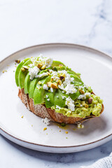 Avocado toast with feta and pistachio on plate, white marble background.