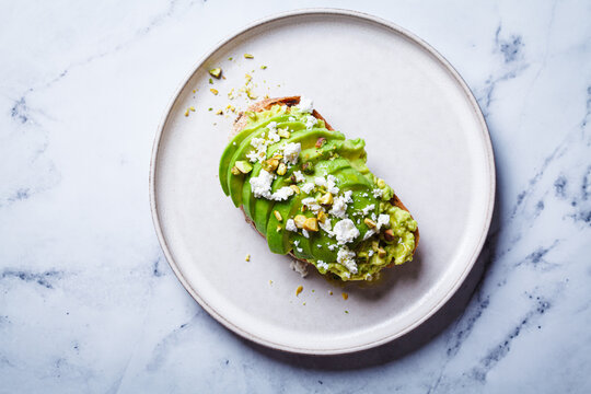 Avocado toast with feta and pistachio on plate, white marble background.
