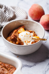 Portion of peach cobbler with scoop of ice cream in bowl, white marble background.