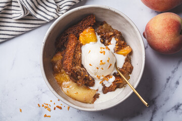 Portion of peach cobbler with scoop of ice cream in bowl, white marble background.