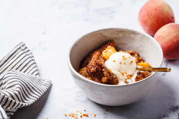 Portion of peach cobbler with scoop of ice cream in bowl, white marble background.
