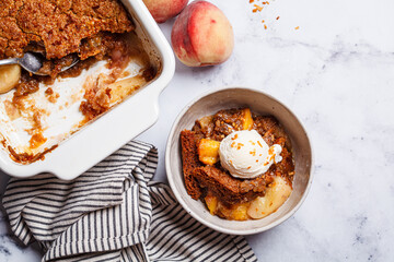 Portion of peach cobbler with scoop of ice cream in bowl, white marble background.