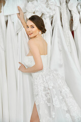 A young, beautiful bride with brunette hair stands in a wedding salon, gazing at a rack of exquisite white dresses.