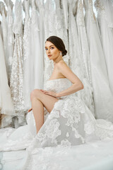 A young brunette bride in a white wedding dress sits gracefully on a bed, contemplating the...