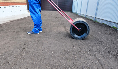 male worker in a uniform prepares a plot of land for sowing a lawn using a garden roller, leveling...