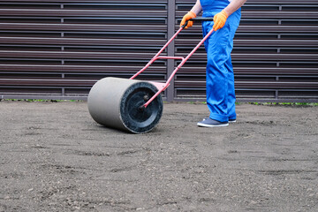 male worker in a uniform prepares a plot of land for sowing a lawn using a garden roller, leveling...