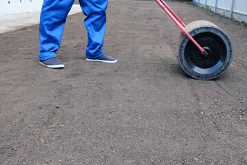 male worker in a uniform prepares a plot of land for sowing a lawn using a garden roller, leveling...