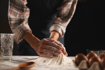 Baker's hands forming a pile of flour on table, plaid shirt, dark background