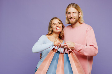 Young smiling amazed couple two friends family man woman wearing pink blue casual clothes hold open shopping paper package bags isolated on plain purple background. Black Friday sale buy day concept.