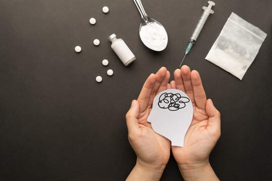 Drug addiction induced psychosis. Hands holding human brain feeling confused symbol with drug powder, syringe and pills on dark background. International Day against Drug Abuse.