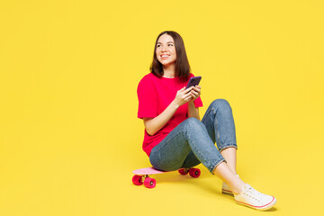 Full body young woman wear pink t-shirt casual clothes sits on skateboard pennyboard hold in hand use mobile cell phone look aside on area isolated on plain yellow orange background Lifestyle concept © ViDi Studio