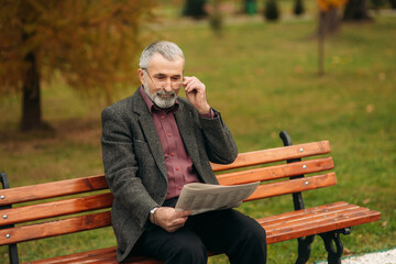 A nice grandfather with a beautiful beard in a gray jacket sits on a bench in the park and reads a newspaper