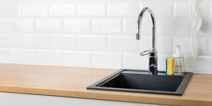 A clean empty black kitchen sink in a kitchen with a chrome faucet, wooden countertop, and white subway tile backsplash with dishwashing liquid, brush and sponge, banner with copy space