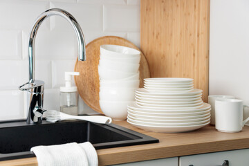 Set of clean washed white plates, tableware near black kitchen sink with a chrome faucet on wooden...
