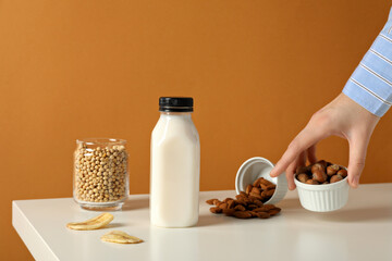 Bottle of milk, hand, nuts in bowls, slices of banana and soy in glass on orange background