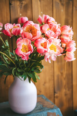 A lovely arrangement of pink peonies in a pink vase, contrasted by a wooden background