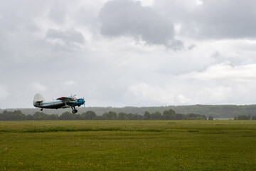 An airplane takes off from a dirt airfield. A biplane flying at low altitude over a field. Cloudy rainy weather. Clouds in the sky. Regional small aviation. An airplane flight in the countryside. © Andrei Stepanov