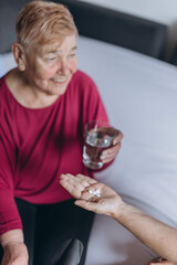 An elderly woman sits on her bed and a volunteer gives her pills and a glass of water
