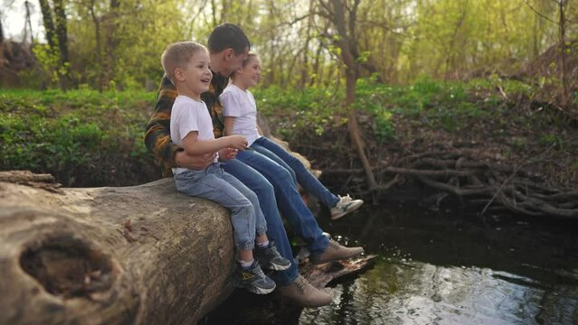 dad with children sits on a log. happy family child dream concept. happy young dad sitting with his son and daughter in the forest on a large log above lifestyle the river. kids dad's