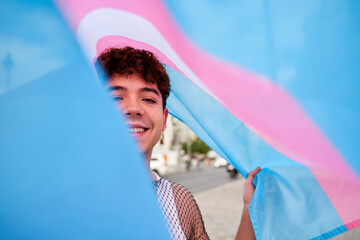 A man is smiling and holding a trans flag. The flag is behind him, and he is wearing a white shirt